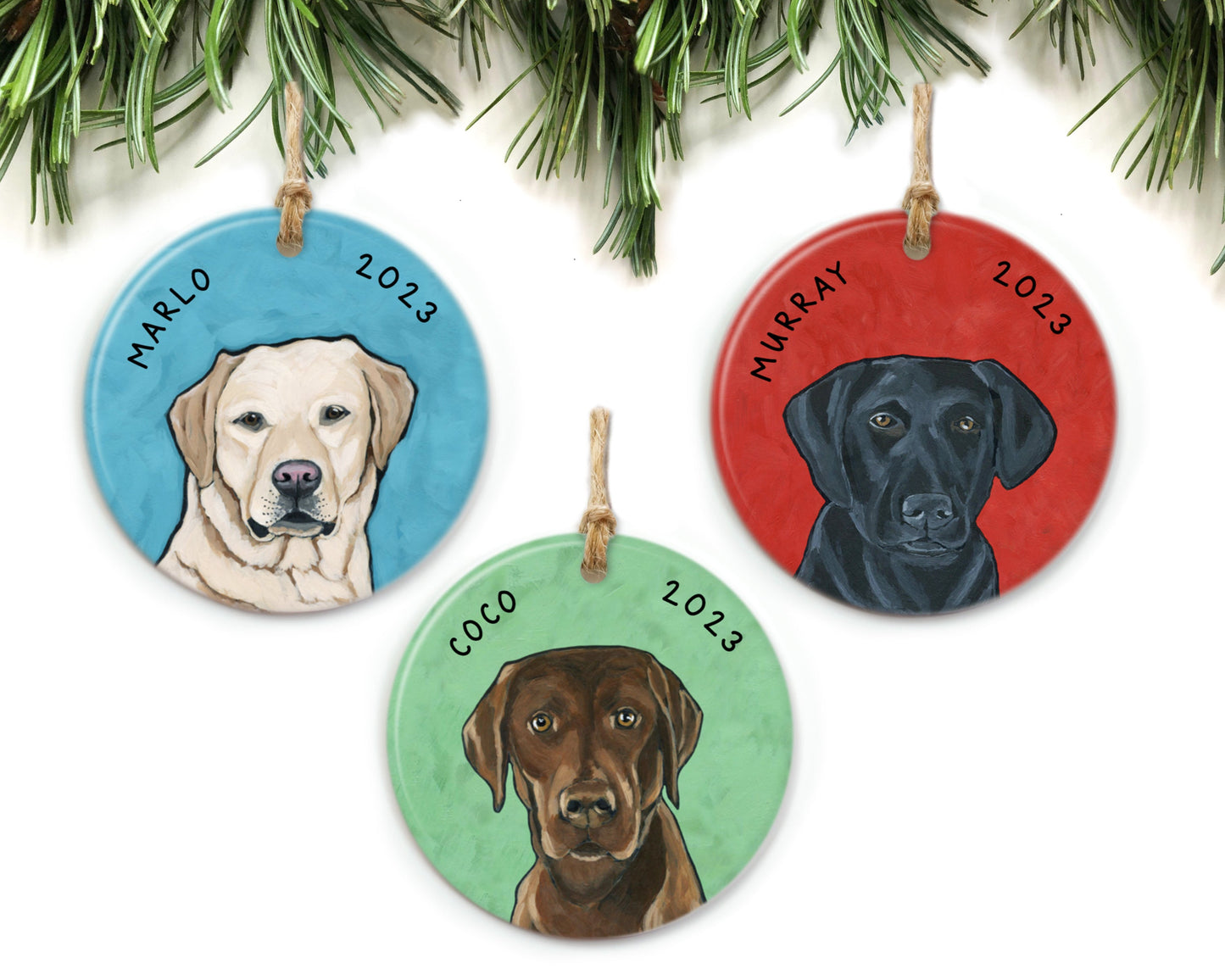 Three dog ornaments hanging from pine branches. Each ornament features a different Labrador Retriever.