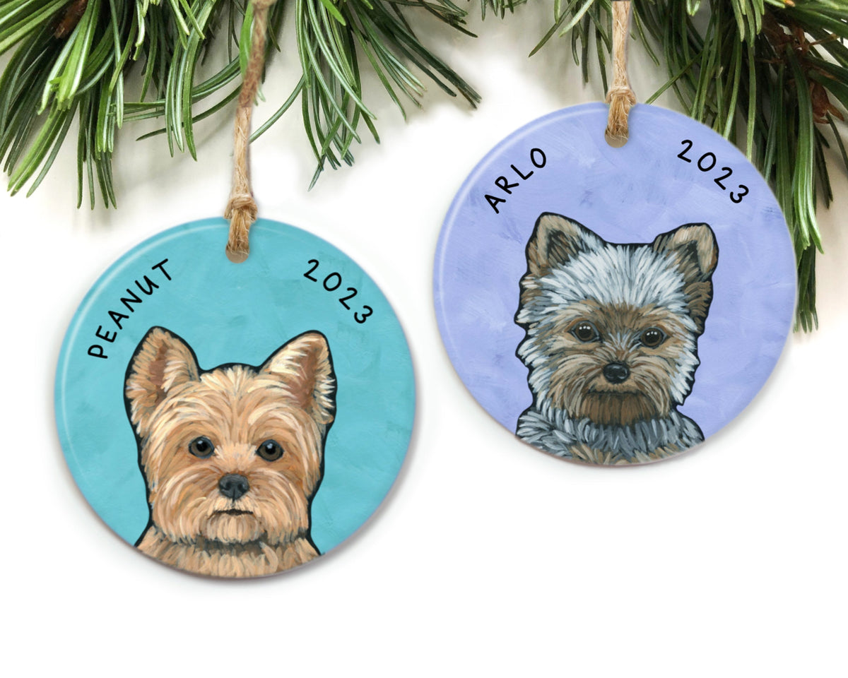 Two Yorkie ornaments by The Cheery Pet (Jessi McNeal) hanging from a pine branch