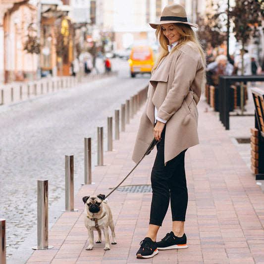 Woman in a beige coat and hat walking a pug on a leash on a city sidewalk